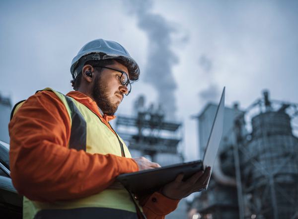 An employee in hardhat and hi-vis jacket works on a laptop outside a natural gas power plant