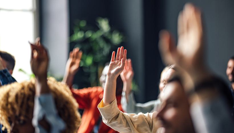 A group of diverse people facing the front of a room raising their hands