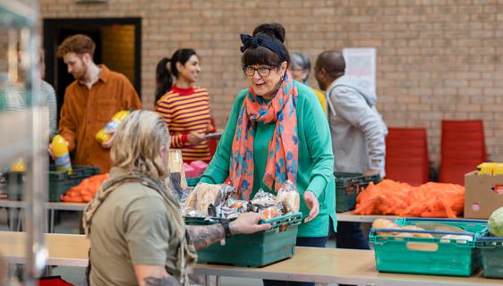 A woman in a black hairband and a green top speaks to a man in a wheelchair over a crate of food