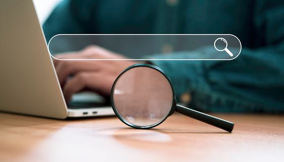 A magnifying glass and search bar on a desk in front of a man's hands working on a laptop