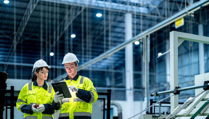 Two employees in hi-vis and hard hats stand in a factory, viewing a tablet computer