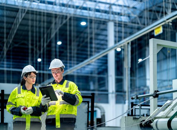 Two employees in hi-vis and hard hats stand in a factory, viewing a tablet computer