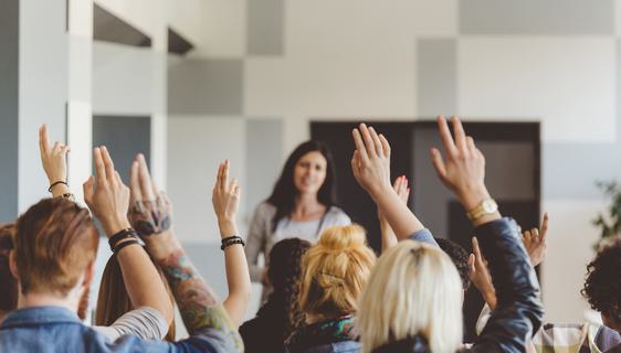 A woman talks to a room of people, many of whom are raising their hands