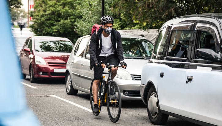 A cyclist rides past a queue of cars, wearing a facemask