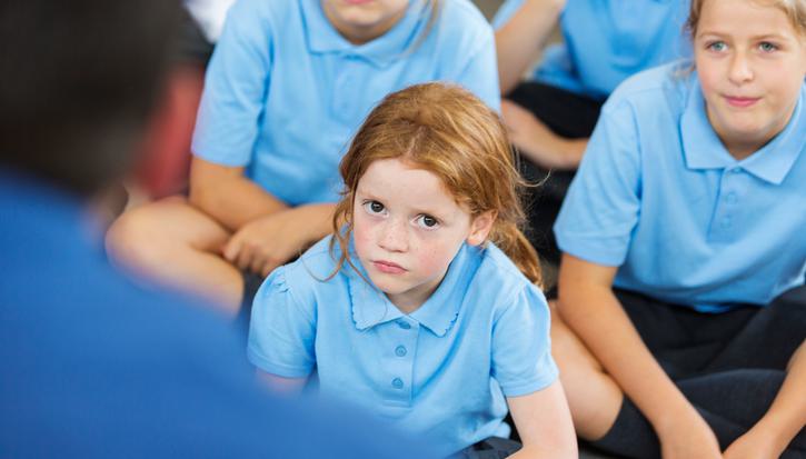 A primary school pupil sits cross-legged in a class, looking at the camera