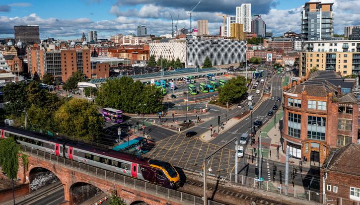 A high-up view of Leeds city centre: a bustling city with a train over a viaduct in the goreground and the city and lots of green buses behind