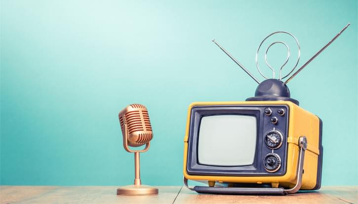 An old fashioned yellow TV and standing microphone sit on a desk in front of a turquoise background