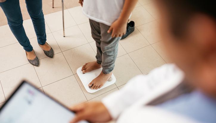 A person stands on a weighing scale, overlooked by a doctor holding a tablet computer